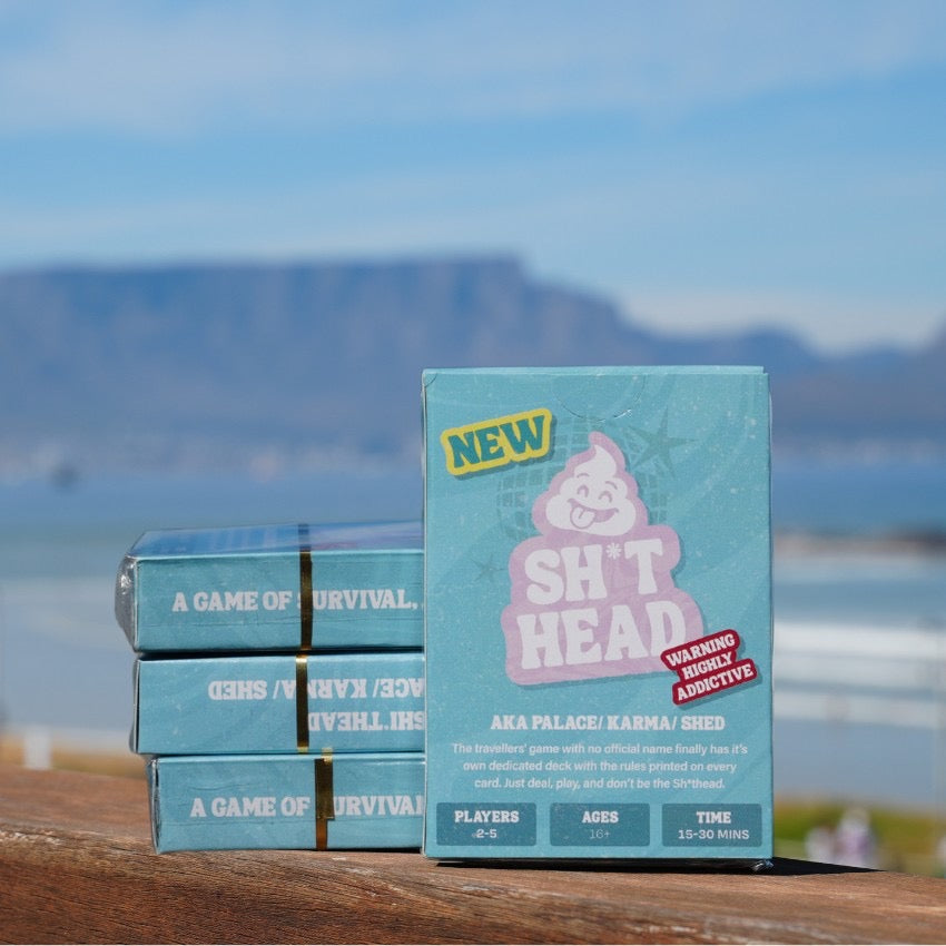 Sh*thead Card Game teal boxes on wooden railing with Table Mountain and sea backdrop