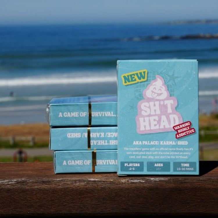 Sh*thead Card Game boxes on a wooden railing overlooking the beach