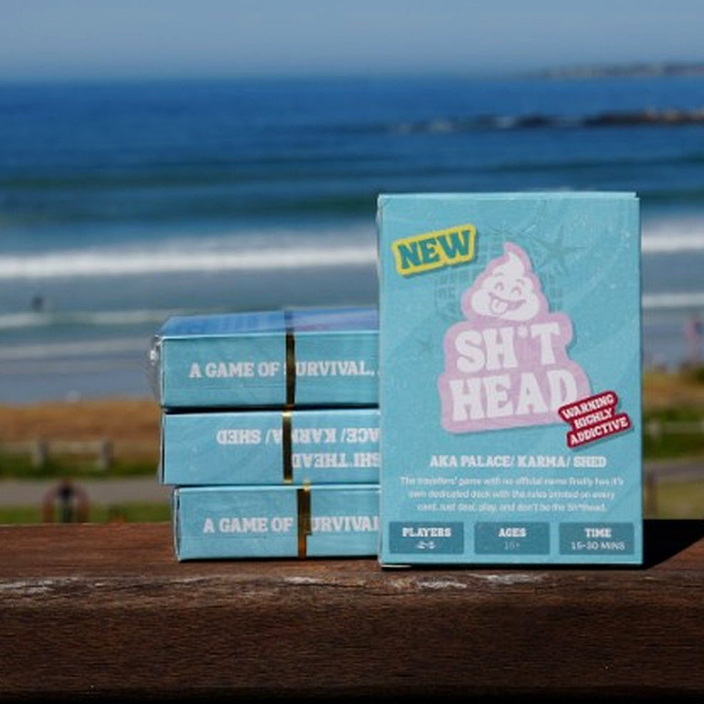 Sh*thead Card Game boxes on a wooden railing overlooking the beach